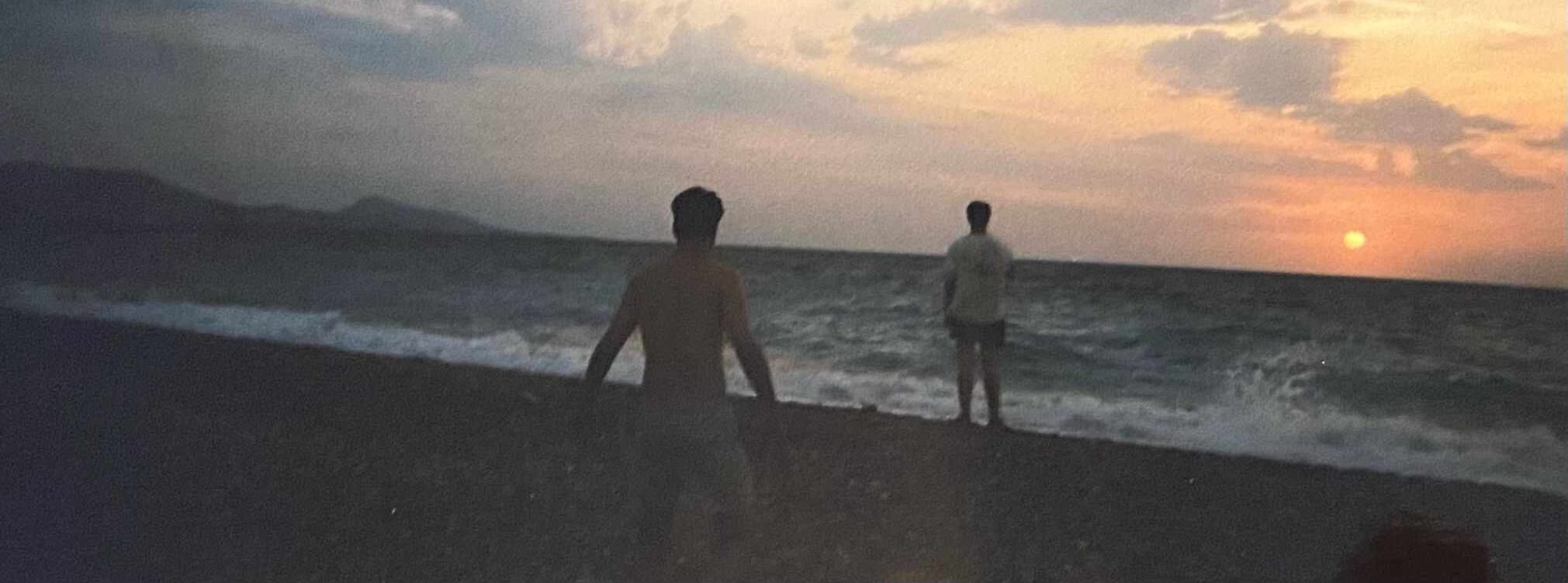 Twee personen staan aan de kust terwijl ze naar de zonsondergang kijken. De zon zakt dicht bij de horizon, kleurend de hemel in tinten van oranje en roze. Golven breken zachtjes op het strand. De contouren van bergen zijn zichtbaar in de verte.